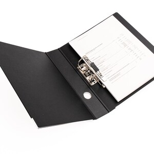 May include: A black binder with a silver metal ring mechanism. The binder is open and has a white sheet of paper inserted.