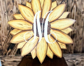 May include: A wooden sunflower door hanger with a white letter "M" in the center. The sunflower is painted yellow with brown accents and has a brown center with white dots.