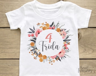 May include: White t-shirt featuring a floral wreath design. The wreath is composed of pink, orange and yellow flowers. In the centre of the wreath is a circle with the number "4" and the name "Frida" written in grey.