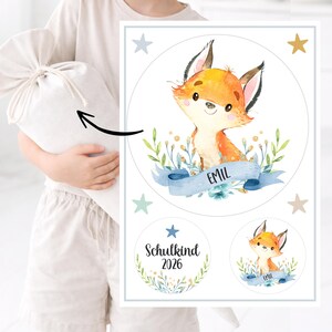 Könnte beinhalten: Aquarell-Illustration eines niedlichen Fuchses mit orange-weißem Fell, umgeben von Grün und einem blauen Band mit dem Namen "EMIL". Das Design beinhaltet Sterne und den Text "Schulkind 2026" in einem kreisförmigen Kranz.