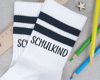 Può includere: Un paio di calzini bianchi con strisce nere e la scritta "SCHULKIND" stampata in nero. I calzini sono accanto a un righello di legno, una stella di legno e tre matite colorate.