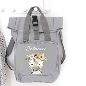 Könnte beinhalten: Ein grauer Rucksack mit Rollverschluss und einer schwarzen Schnalle. Die Vorderseite des Rucksacks zeigt ein weißes Zebra mit einer Blumenkrone und den Namen "Antonia" in weiß.