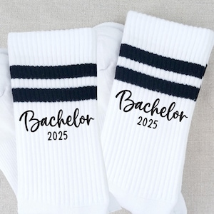 Könnte beinhalten: Paar weiße Sportsocken mit marineblauen Streifen und dem Wort "Bachelor" und dem Jahr "2025" in schwarzer Schrift. Die Socken sind gerippt und haben ein sportliches Design.