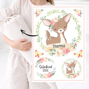 Könnte beinhalten: Ein hellfarbener Beutel mit passender Illustration eines Rehs mit Blumenkrone, umgeben von einem Kranz aus Blättern und Blumen. Der Name "Theresa" steht auf einem Banner. Zusätzliche kreisförmige Designs zeigen ein Reh und den Text "Schulkind 2026".