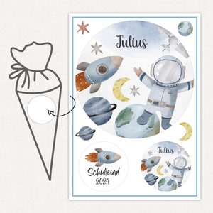 Könnte beinhalten: Aquarellillustration eines Astronauten, Planeten, Raketen und Sterne auf weißem Hintergrund. Der Name "Julius" ist in Schreibschrift geschrieben. Ein Aufkleber mit dem Text "Schulkind 2024" ist ebenfalls enthalten.