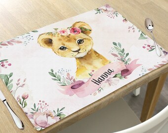 May include: A white placemat with a watercolor illustration of a lion cub wearing a flower crown. The placemat has pink and green floral accents and the name "Hanna" written in black script.
