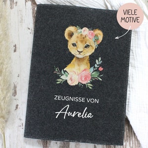 Könnte beinhalten: Ein dunkelgrauer Filzordner mit einer Löwenjungendarstellung. Das Löwenjungtier trägt eine rosafarbene Blumenkrone und ist von rosa und weißen Blumen umgeben. Der Text "Zeugnisse von Aurelia" ist auf dem Ordner aufgedruckt.