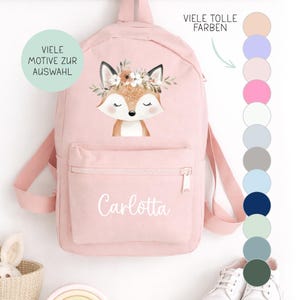 Könnte beinhalten: Ein hellrosa Rucksack mit einer Vordertasche und Reißverschluss. Der Rucksack zeigt eine Fuchsgrafik mit einem Blumenkranz und dem Namen "Carlotta" in weißer Schrift. Der Text "VIELE MOTIVE ZUR AUSWAHL" und "VIELE TOLLE FARBEN" sind ebenfalls sichtbar.