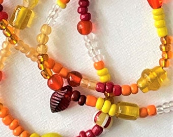 May include: A multi-strand beaded necklace with orange, yellow, red, and clear beads. The necklace features a variety of bead shapes and sizes, including round, square, and leaf-shaped beads.