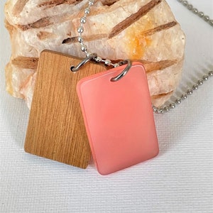 May include: A silver chain necklace with two rectangular pendants. One pendant is made of light brown wood and the other is made of translucent pink resin.