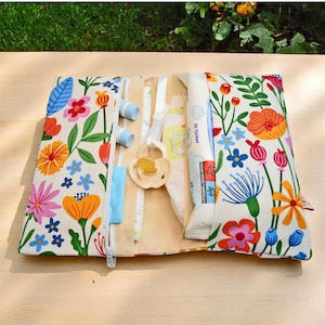 Könnte beinhalten: Eine weiße Reißverschlusstasche mit einem Blumendruck in Rosa-, Blau-, Gelb- und Orangetönen. Die Tasche ist geöffnet und zeigt eine Packung Babyfeuchttücher, einen Holz-Beißring und ein paar Babyfeuchttücher.