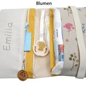 May include: A white zippered pouch with a yellow and white polka dot fabric lining. The pouch is open and contains a pacifier, a package of baby wipes, and a small wooden mushroom charm. The pouch is embroidered with the name "Emilia" and has a floral print on the outside.