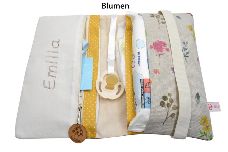 May include: A white zippered pouch with a yellow and white polka dot fabric lining. The pouch is open and contains a pacifier, a package of baby wipes, and a small wooden mushroom charm. The pouch is embroidered with the name "Emilia" and has a floral print on the outside.