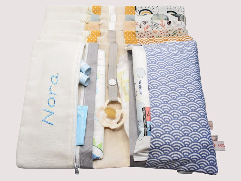 May include: A white zippered pouch with the name "Nora" embroidered on the front. The pouch is open to reveal several compartments, including a blue and white patterned fabric, a white fabric with a pacifier, and a white fabric with the text "30 T&uuml;cher".