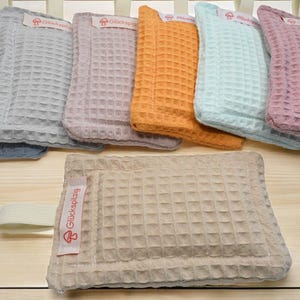 Könnte beinhalten: Ein Set aus acht Waffelgewebe-Waschlappen in verschiedenen Farben, darunter Blau, Grau, Rosa, Beige und Orange. Jeder Waschlappen hat ein kleines Etikett mit dem Text "Glücksplag" in Rot.