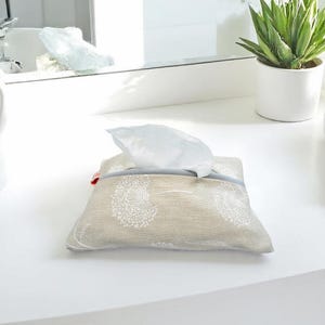 Könnte beinhalten: Eine beige Stoff-Taschentuchbox-Hülle mit einem weißen Pusteblumenmuster. Die Hülle hat eine graue Einfassung und einen roten Akzent. Weiße Taschentücher werden aus der oberen Öffnung gezogen. Die Taschentuchbox-Hülle befindet sich auf einer weißen Oberfläche.