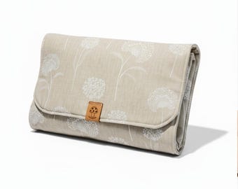 Puede incluir: Una alfombrilla rectangular plegada de tela beige con un estampado de dientes de león blancos. La alfombrilla tiene una pequeña lengüeta de cuero marrón centrada en la solapa frontal, grabada con un dibujo lineal de un hongo con sombrero punteado.