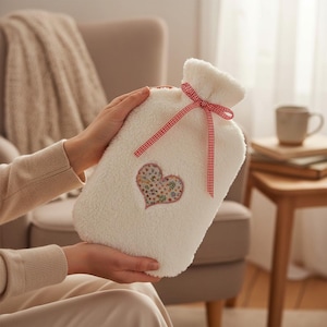 Könnte beinhalten: Cremefarbene Wärmflasche mit weichem, plüschigem Bezug. Sie hat ein herzförmiges Blumen-Applikation und ein rot-weiß kariertes Band. Das Design deutet auf ein gemütliches und komfortables Produkt hin.