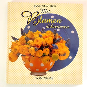 Peut inclure: Un livre intitulé "Mit Blumen dekorieren" de Jane Newdick. La couverture présente un bol à pois jaunes et orange rempli de fleurs jaunes et de brins bleus. Le bol est posé sur une assiette bleue à pois blancs. L'arrière-plan est un motif jaune à pois blancs.