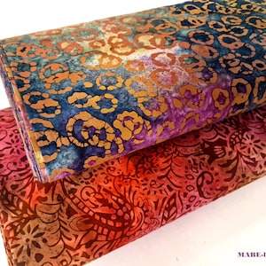 Op de afbeelding: Twee stukken stof met een batikpatroon. De bovenste stof is een blauwe, paarse en gouden batik met een cirkelvormig patroon. De onderste stof is een rode, oranje en bruine batik met een bloemmotief.