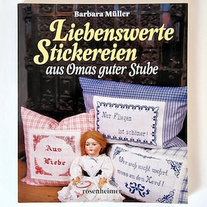Könnte beinhalten: Ein Buchcover mit einer Vintage-Illustration von Kissen mit gestickten Sprüchen auf Deutsch. Der Buchtitel lautet "Liebenswerte Stickereien aus Omas guter Stube", was übersetzt "Liebenswerte Stickereien aus Omas guter Stube" bedeutet.