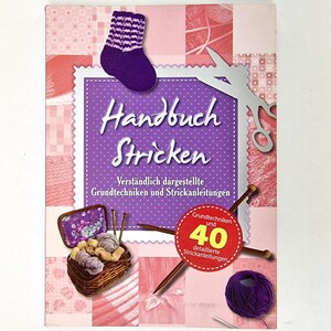 Könnte beinhalten: Ein lilafarbenes Buch mit dem Titel "Handbuch Stricken" und einer lilafarbenen Socke auf dem Cover. Das Buch hat einen rosa-weiß karierten Hintergrund mit einem Korb voller Wolle und Stricknadeln. Der Text auf dem Cover lautet "Verständlich dargestellte Grundtechniken und Strickanleitungen" und "Grundtechniken und 40 detaillierte Strickanleitungen".