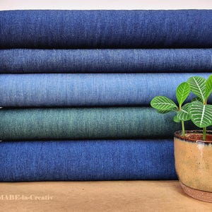 Może przedstawiać: Stos rolek materiału dżinsowego w różnych odcieniach niebieskiego i turkusowego. Rolki są starannie ułożone, prezentując różne przejścia kolorystyczne. Mała roślina doniczkowa z zielonymi liśćmi stoi przed tkaniną.