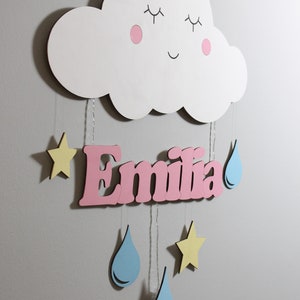 May include: A white wooden cloud with a smiling face, pink cheeks, and closed eyes hangs from the ceiling.  The cloud is attached to a pink wooden name sign that reads "Emilia".  The name sign is decorated with yellow stars and blue raindrops.
