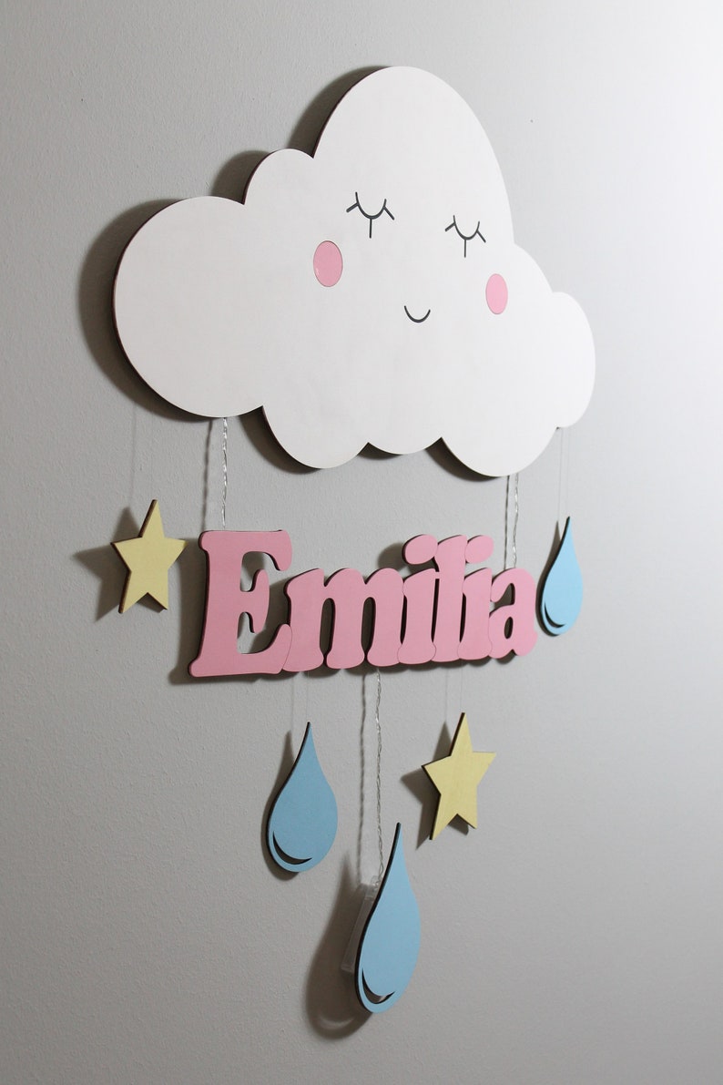 May include: A white wooden cloud with a smiling face, pink cheeks, and closed eyes hangs from the ceiling.  The cloud is attached to a pink wooden name sign that reads "Emilia".  The name sign is decorated with yellow stars and blue raindrops.