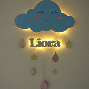 May include: A blue wooden cloud with a smiling face and closed eyes, with a name "Liora" written in brown wood below. The cloud and name are illuminated with warm white lights. Hanging from the cloud are pink and yellow wooden raindrops and stars.