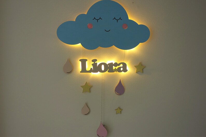 May include: A blue wooden cloud with a smiling face and closed eyes, with a name "Liora" written in brown wood below. The cloud and name are illuminated with warm white lights. Hanging from the cloud are pink and yellow wooden raindrops and stars.