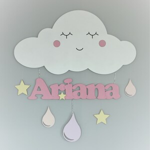 May include: A pink wooden cloud with a smiling face and closed eyes, with the name "Ariana" written in pink letters below. The cloud has pink cheeks and is decorated with yellow stars and pink raindrops.