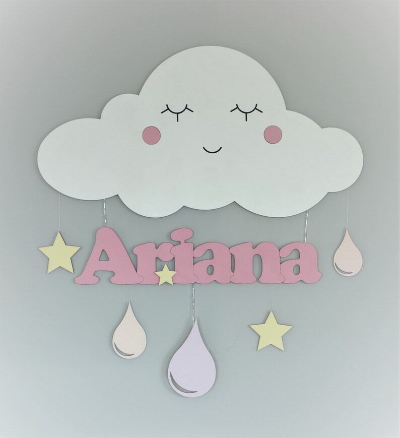 May include: A pink wooden cloud with a smiling face and closed eyes, with the name "Ariana" written in pink letters below. The cloud has pink cheeks and is decorated with yellow stars and pink raindrops.