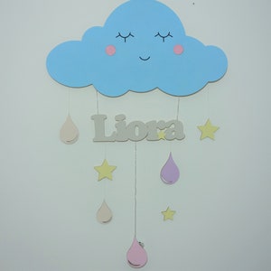 May include: A blue cloud with a smiling face and pink cheeks hangs from the ceiling with a name sign that reads "Liora" and several beige and pink raindrops and yellow stars hanging below.