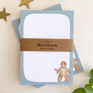Könnte beinhalten: Hellblaue Notizblöcke mit abgerundeten Ecken, zusammengehalten von einem braunen Band. Der Notizblock zeigt eine Lebkuchenmann-Illustration mit einer Zuckerstange. Der Text "ohl happy paper Notizblock DIN A6, 50 Blatt" ist auf dem Band aufgedruckt.