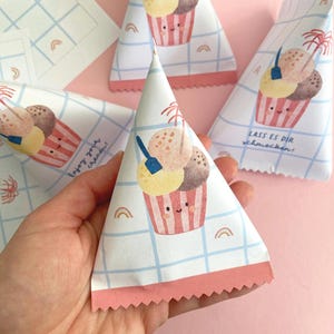 May include: Triangular paper treat bags with a cartoon ice cream cone design. The bags feature a white background with a blue grid pattern, rainbows, and the words "Enjoy your cream!" in black. The ice cream cones have smiling faces.