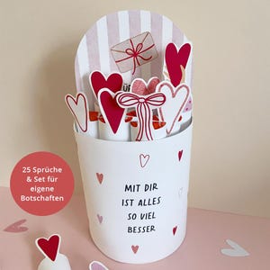 May include: A white container with red hearts and the text "MIT DIR IST ALLES SO VIEL BESSER". The container holds several heart-shaped decorations and a paper envelope with a red bow. The background has pink and white stripes.
