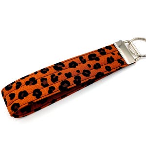 Può includere: Un portachiavi in tessuto color arancione bruciato con un motivo leopardato nero. Il portachiavi ha una chiusura e un portachiavi in metallo color argento. Il tessuto è a coste e il portachiavi è leggermente inclinato verso destra.