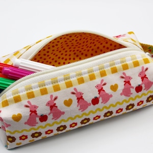 May include: A white fabric pencil case with a yellow and white checkered pattern and a pink bunny rabbit design. The case has a zipper closure and a pink fabric handle.