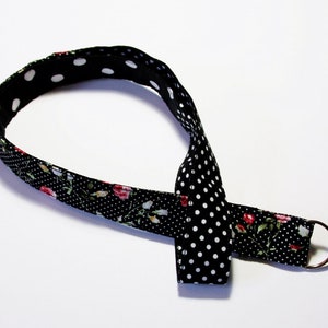 Könnte beinhalten: Schwarzes Stoffband mit weißen Punkten und einem floralen Muster auf der anderen Seite. Das Band hat einen silbernen Metallring am Ende.