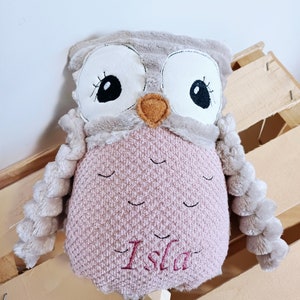 Peut inclure: Un jouet en peluche hibou rose et gris avec des yeux brodés et un bec. Le hibou a une longue queue tressée et le nom "Isla" est brodé sur sa poitrine.