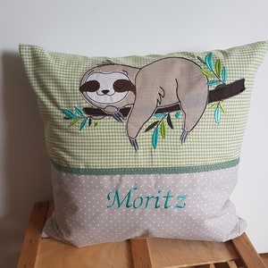 May include: A green and beige pillow with a brown sloth embroidered on it. The sloth is sleeping on a branch with green leaves. The pillow has a green and white gingham pattern on the top half and a beige polka dot pattern on the bottom half. The name "Moritz" is embroidered in green on the bottom half of the pillow.