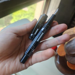 May include: A black fountain pen with a geometric white design is held in a hand. The pen's cap is off, revealing the nib. The pen has a sleek, modern design, with a black body and silver-colored nib.