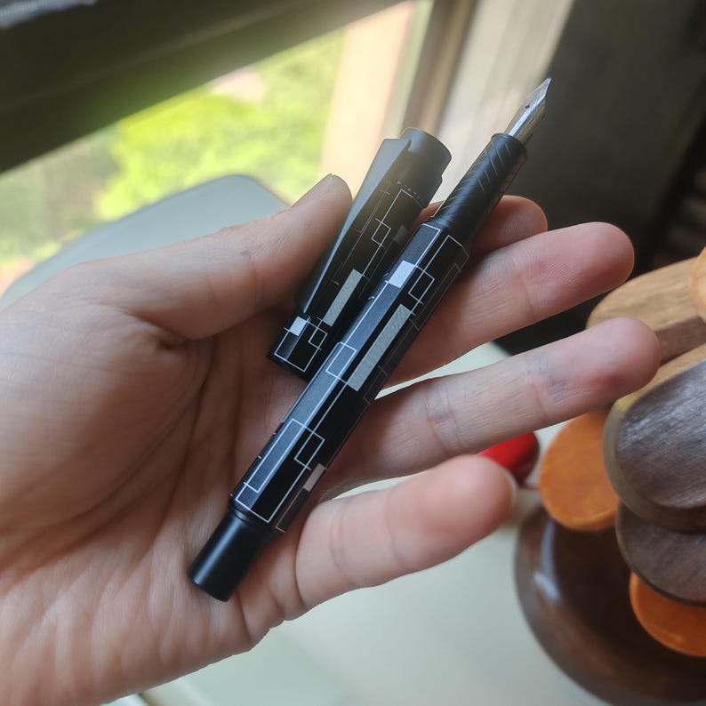 May include: A black fountain pen with a geometric white design is held in a hand. The pen's cap is off, revealing the nib. The pen has a sleek, modern design, with a black body and silver-colored nib.