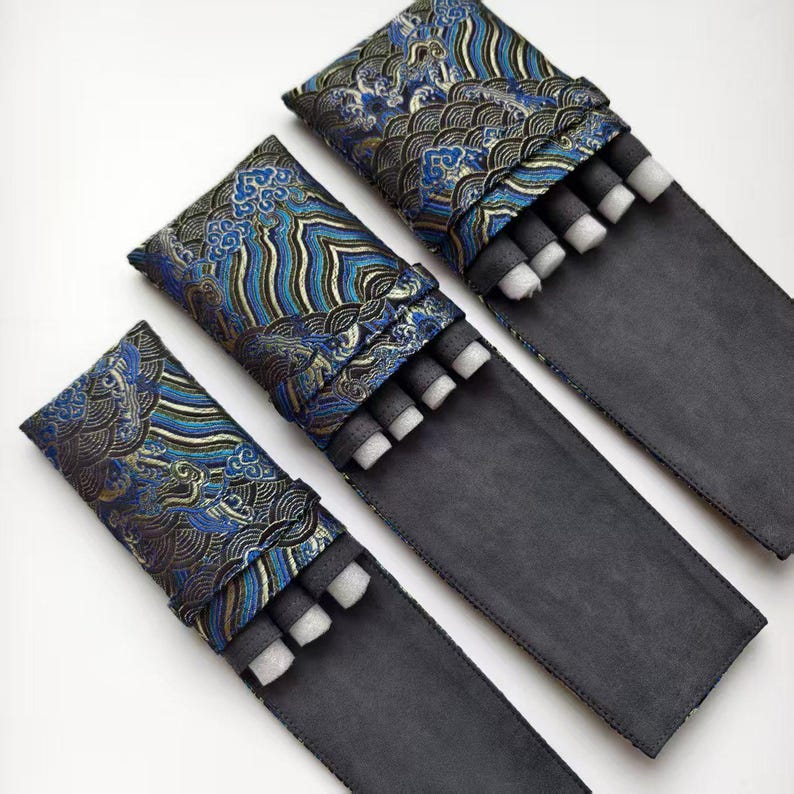 May include: Three black and blue fabric pen cases with decorative patterned tops. Each case holds several black pens with white foam inserts. The cases are arranged on a white surface.