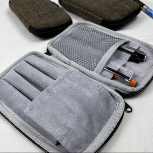 May include: Open gray pen case with a black zipper, revealing a gray interior with pen slots and a mesh pocket. Two additional cases with a brown tweed exterior are visible in the background. Pens are inside the open case.