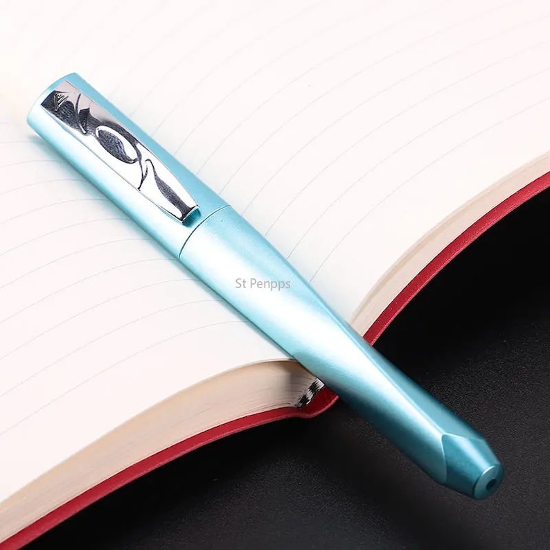 May include: A light blue fountain pen with a silver trim and a silver design on the cap. The pen is lying on a notebook with lined pages.