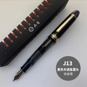 May include: A black and gold fountain pen with a clear ink cartridge. The pen has a gold trim and a black nib. The pen is in a black box with a red and white pattern.