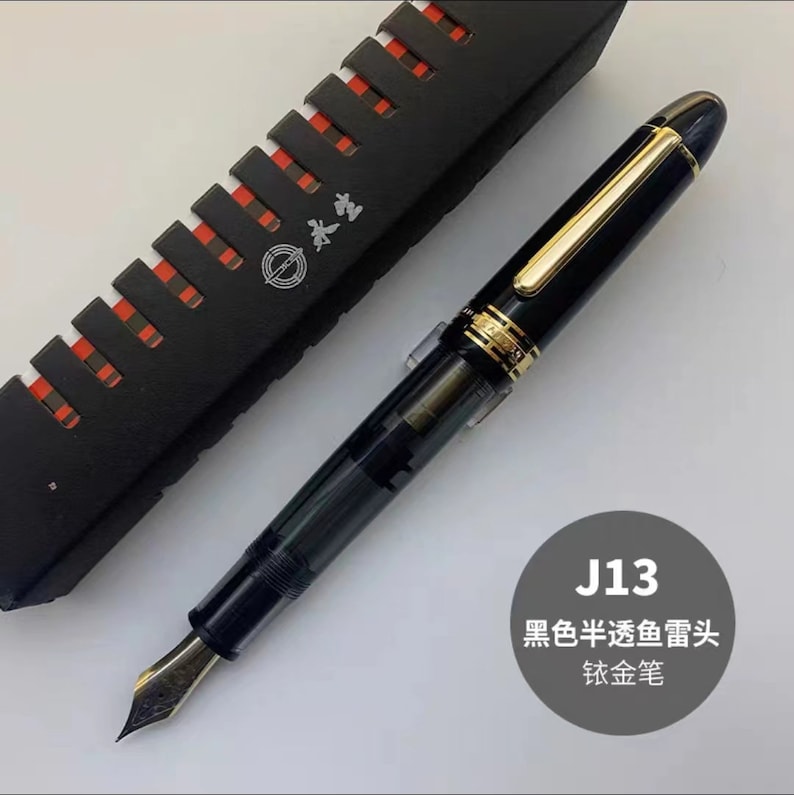 May include: A black and gold fountain pen with a clear ink cartridge. The pen has a gold trim and a black nib. The pen is in a black box with a red and white pattern.
