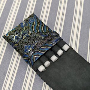 May include: A black pen case with a decorative flap featuring a blue, gold, and black wave pattern. The case holds several black cylindrical objects with white foam inserts, likely for protection. The case is on a striped background.